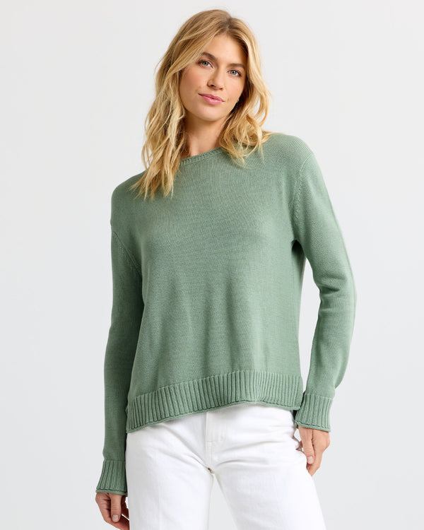 Woman wearing sage green cashmere sweater and white pants against plain light background