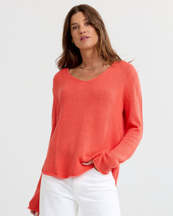 Three-quarter image of a woman wearing coral V-neck cashmere sweater and white pants with hand in pocket