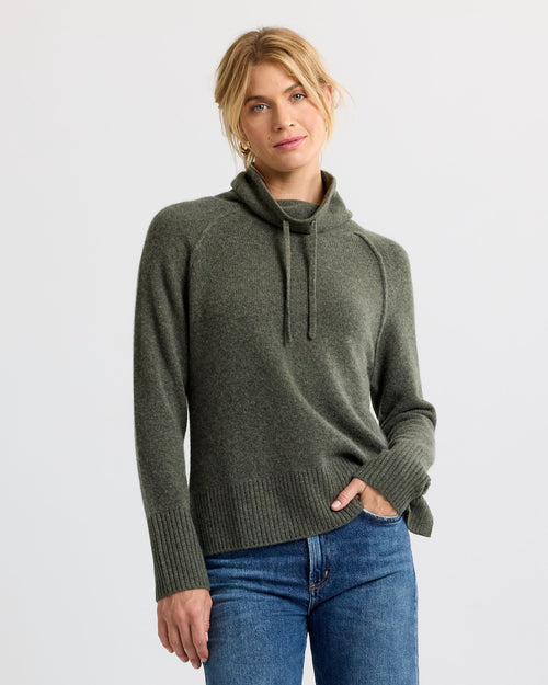 Woman wearing dark olive cashmere sweater with high neck and drawstrings, paired with blue jeans, studio background
