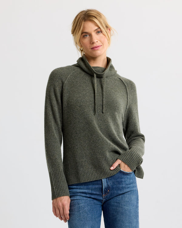 Woman wearing dark olive cashmere sweater with high neck and drawstrings, paired with blue jeans, studio background