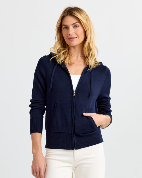 LIFESTYLE / EDITORIAL: Woman wearing navy zip-up cashmere hoodie with pockets and white pants against plain background