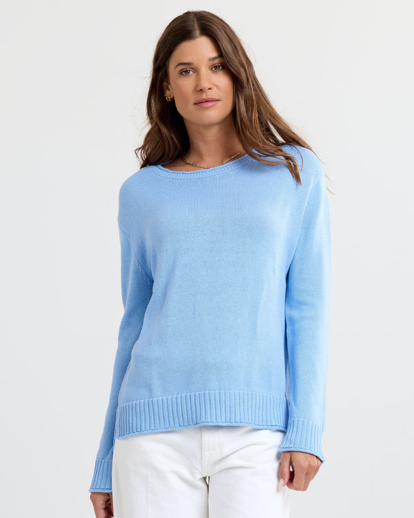 Front-facing image of a woman wearing a light blue cashmere sweater and white pants against a plain background