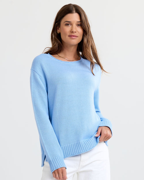 Three-quarter view image of a woman wearing a light blue cashmere sweater and white pants against a plain background