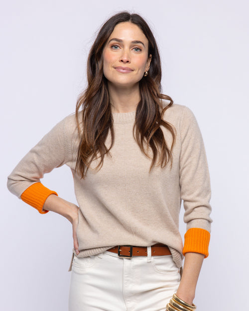 LIFESTYLE / EDITORIAL: Woman wearing beige cashmere sweater with orange cuffs and white pants against light background