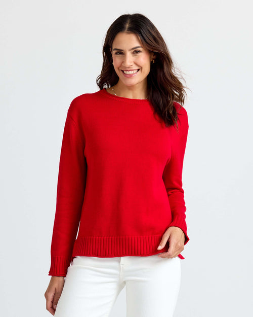 Woman wearing red cashmere sweater and white pants, standing and smiling against white background