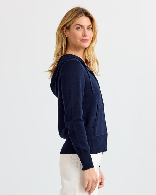LIFESTYLE / EDITORIAL: Woman in navy cashmere hoodie and white pants standing sideways against a plain background