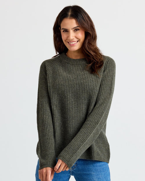LIFESTYLE / EDITORIAL: Woman wearing dark olive ribbed cashmere sweater and blue jeans, smiling against a plain background