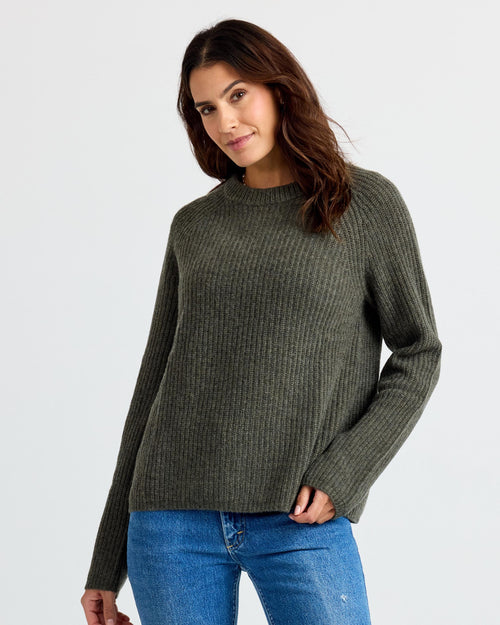 Three-quarter view image of a woman wearing dark olive ribbed cashmere sweater and blue jeans against plain background