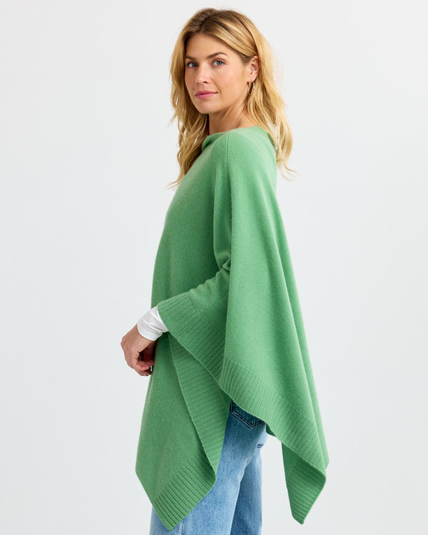 Woman wearing green cashmere poncho over white top and jeans, standing sideways against white background