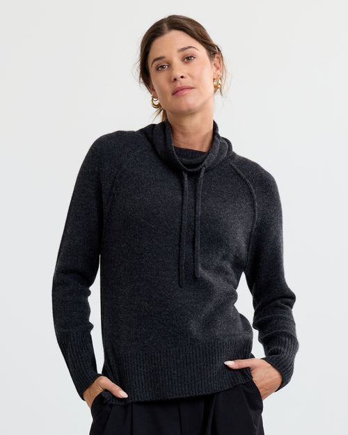 LIFESTYLE / EDITORIAL: Woman wearing charcoal cashmere sweater with cowl neck and drawstrings, hands in pockets, neutral