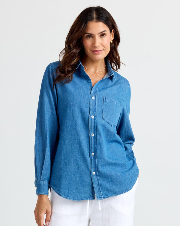 Woman wearing medium wash blue denim shirt with white buttons and chest pocket, standing against plain background