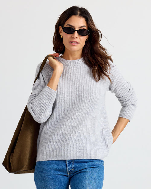 Woman wearing light gray ribbed cashmere sweater, blue jeans, sunglasses, and carrying large brown suede bag
