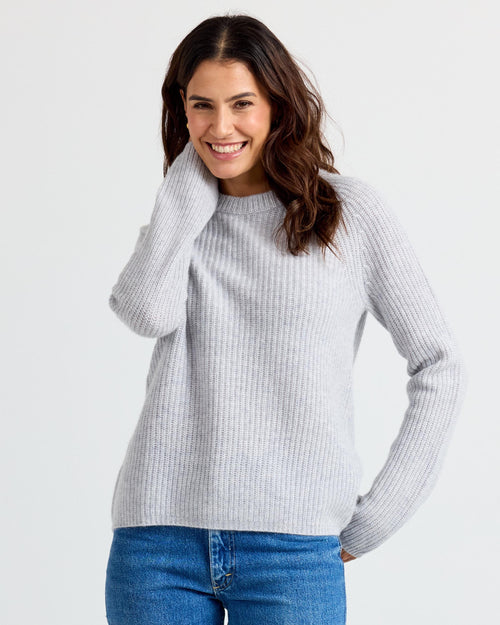 LIFESTYLE / EDITORIAL: Woman wearing a light gray ribbed cashmere sweater and blue jeans, smiling with hand near face