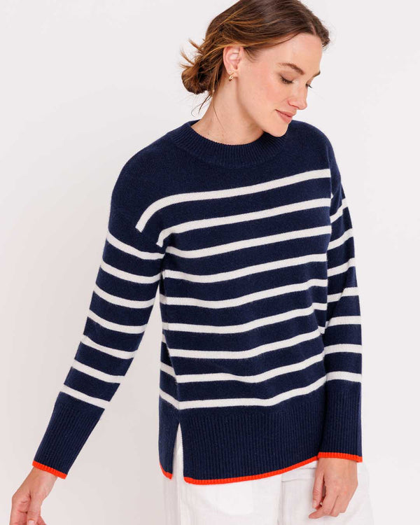 Woman wearing navy and ivory striped cashmere crewneck sweater with orange trim, looking down, white pants visible