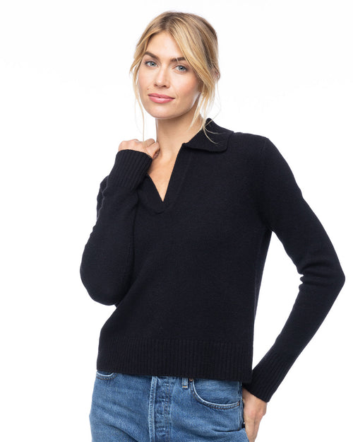 Woman wearing black cashmere polo V-neck sweater and blue jeans, standing against white background