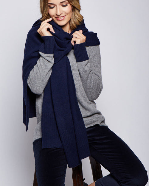 Woman wearing grey sweater and navy ribbed cashmere travel wrap, seated on stool against plain background