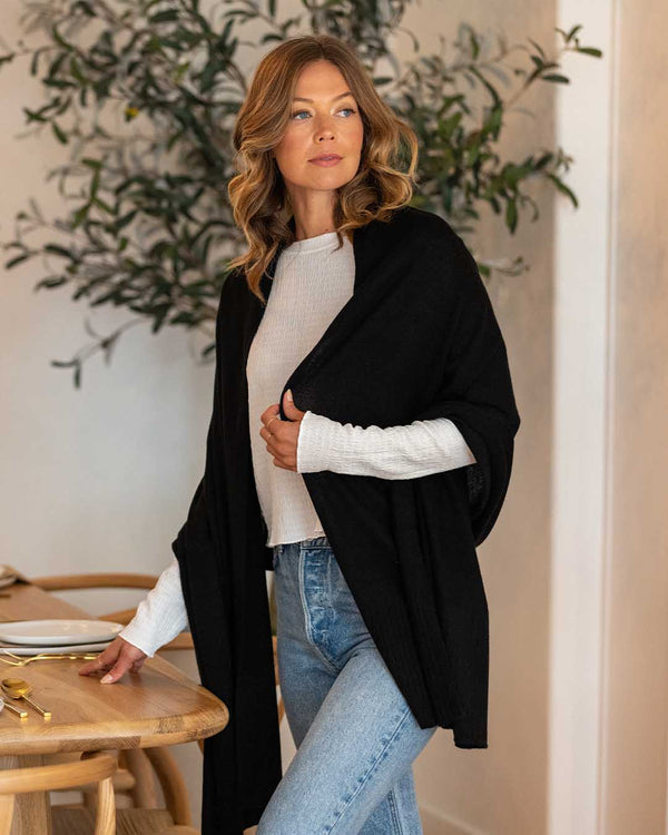 LIFESTYLE / EDITORIAL: Woman wearing black cashmere wrap over white top and jeans, standing by wooden table indoors