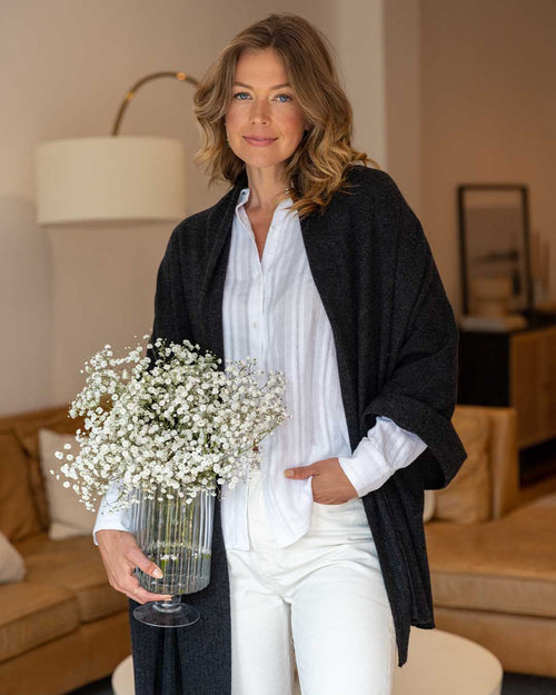LIFESTYLE / EDITORIAL: Woman indoors holding vase with white flowers, wearing charcoal cashmere wrap and white outfit