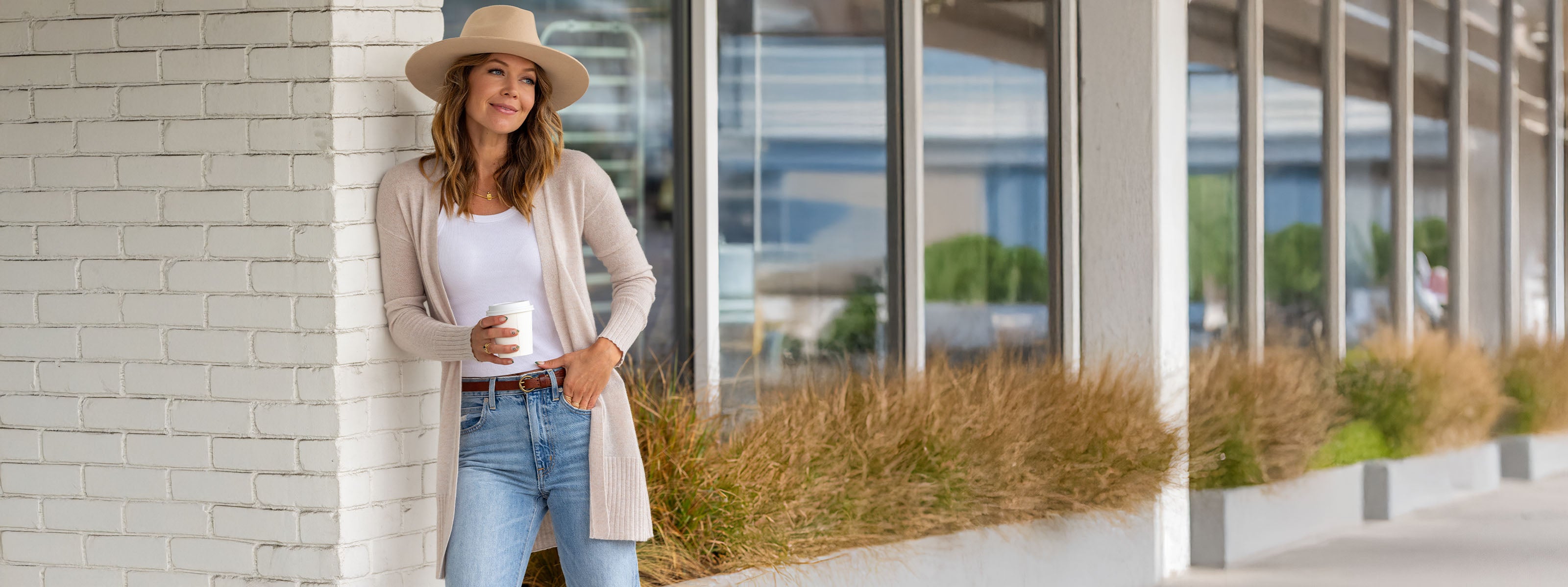 LIFESTYLE / EDITORIAL: Woman in beige cardigan and hat holding coffee, standing by window with plants outside