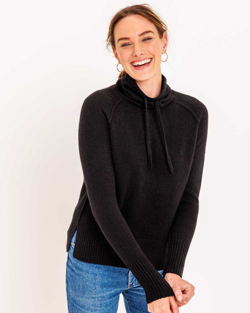 LIFESTYLE / EDITORIAL: Smiling woman wearing black drawstring funnel neck cashmere sweater and blue jeans against white