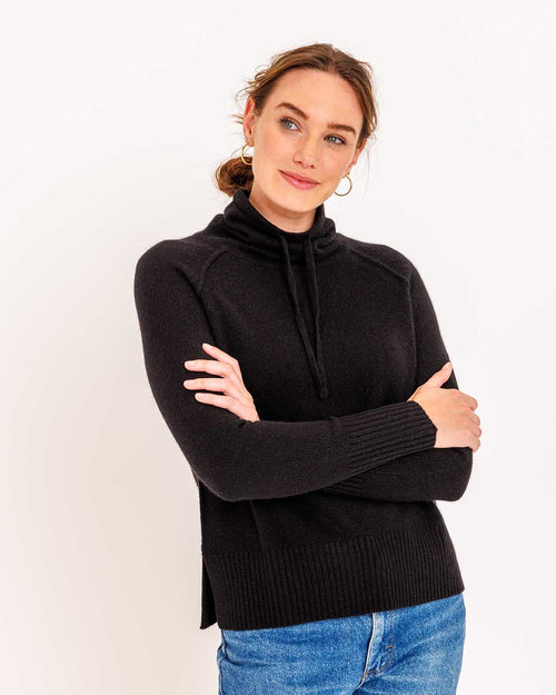 LIFESTYLE / EDITORIAL: Woman wearing black drawstring funnel neck cashmere sweater with blue jeans, arms crossed, white