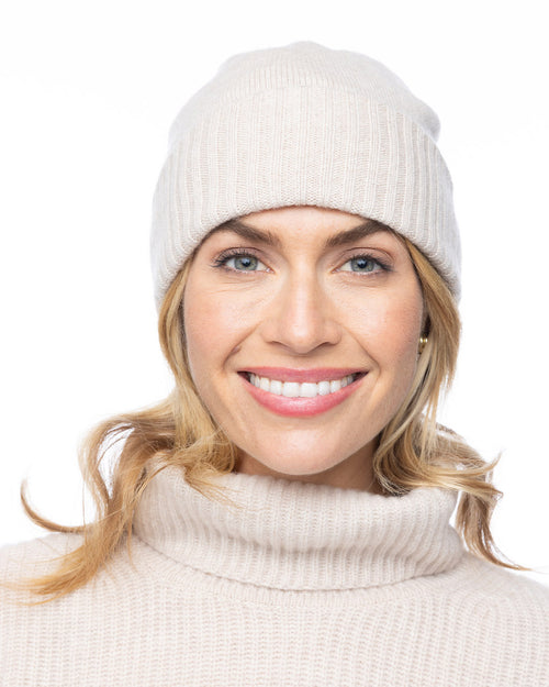 LIFESTYLE / EDITORIAL: Smiling woman wearing oatmeal-colored cashmere hat and turtleneck sweater against white background