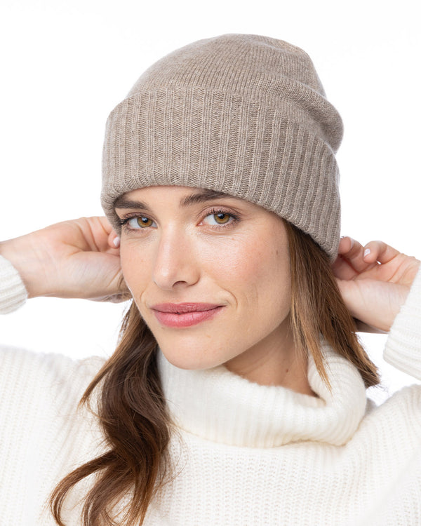 LIFESTYLE / EDITORIAL: Woman wearing beige cashmere beanie and white turtleneck sweater, adjusting hat with hands behind head