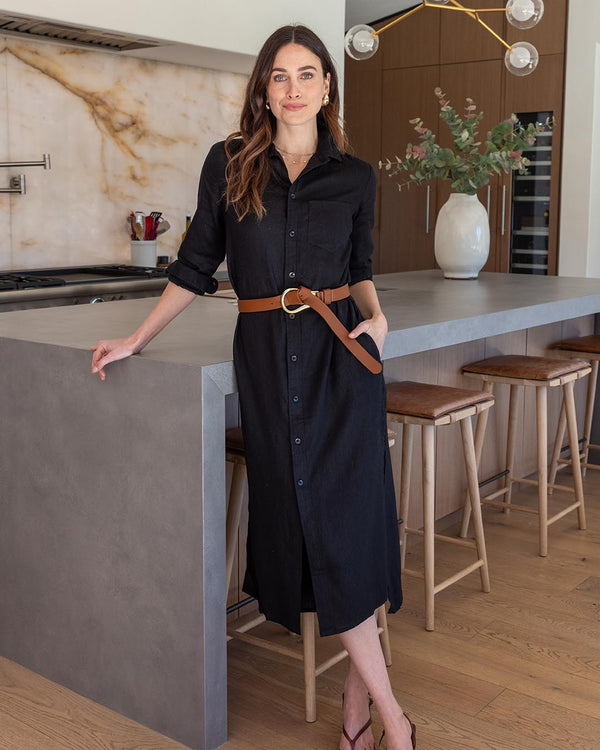 LIFESTYLE / EDITORIAL: Woman in black linen shirt dress with brown belt standing by kitchen island with stools and vase of