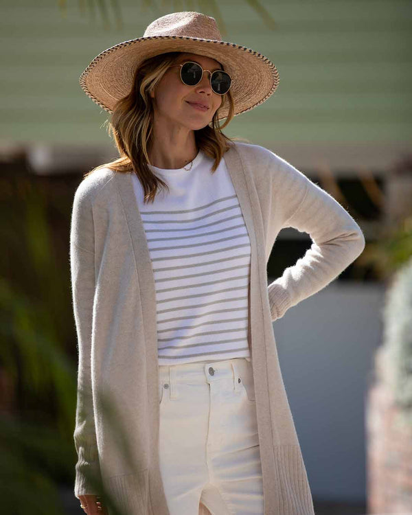 LIFESTYLE / EDITORIAL: Woman wearing oatmeal long cashmere cardigan, striped shirt, white pants, straw hat, and sunglasses