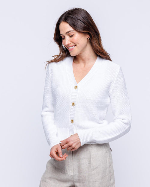Woman wearing white textured cotton cardigan with buttons, smiling and looking down against plain background