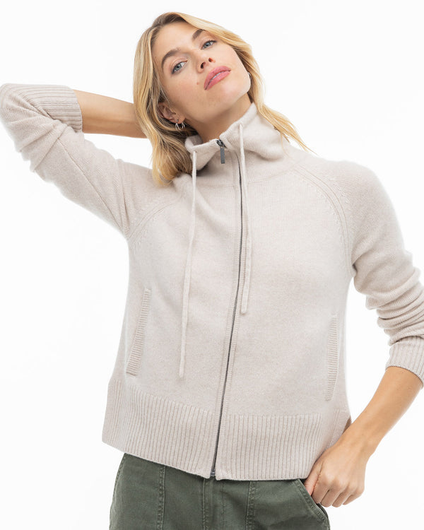 LIFESTYLE / EDITORIAL: Woman wearing oatmeal zip-up cashmere sweater with high collar and drawstrings, posing with hand