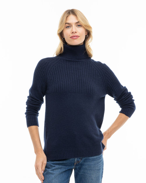 LIFESTYLE / EDITORIAL: Woman wearing navy blue ribbed turtleneck cashmere sweater and jeans, standing against white