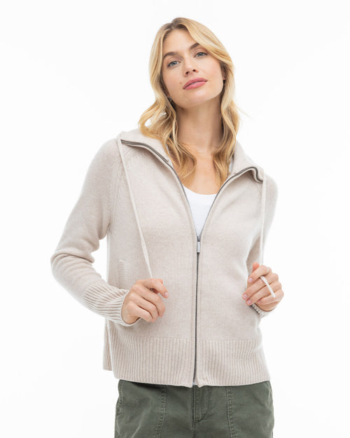 Woman wearing oatmeal zip-up cashmere hoodie over white top, posing against white background
