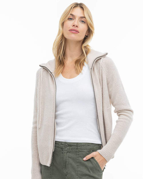 Woman wearing oatmeal zip-up cashmere hoodie, white tank top, and green pants against white background