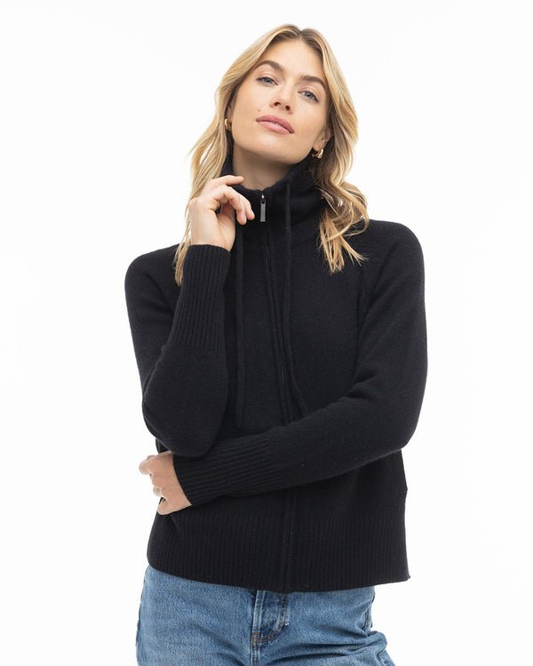 Front-facing image of a woman wearing a black zip-up cashmere sweater and blue jeans with hand near face