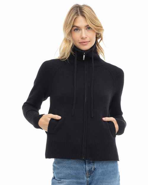 LIFESTYLE / EDITORIAL: Woman wearing black zip-up cashmere sweater with pockets and high collar, standing against white