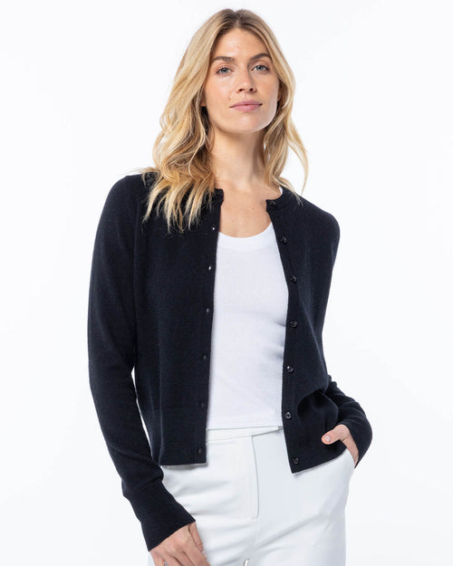 LIFESTYLE / EDITORIAL: Woman wearing black cashmere cardigan over white tank top and white pants, standing against white