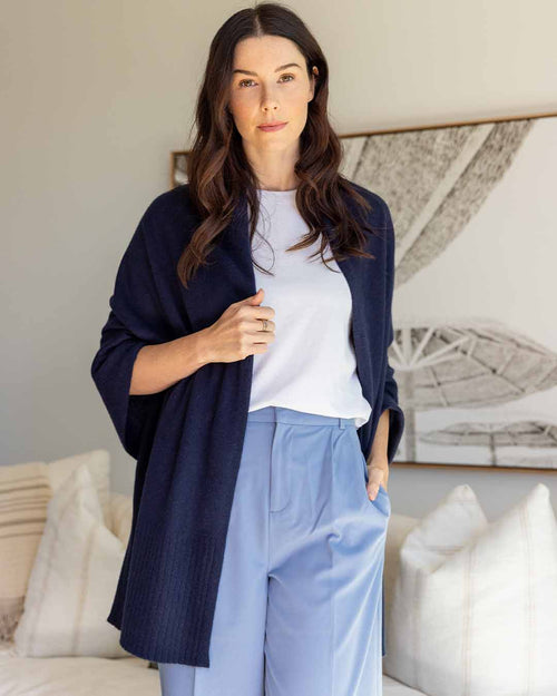 LIFESTYLE / EDITORIAL: Woman indoors wearing navy cashmere wrap over white top and light blue pants, standing by couch and