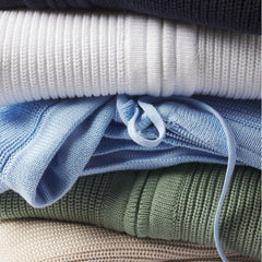 Stack of folded cashmere sweaters in navy, white, light blue, green, and beige, showing knit textures and drawstrings