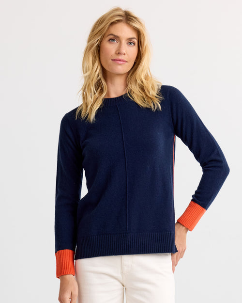 LIFESTYLE / EDITORIAL: Woman wearing navy cashmere sweater with orange cuffs and white pants against white background