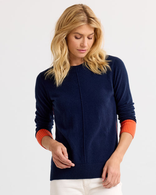 Woman wearing navy blue cashmere sweater with orange cuffs, standing against a plain light background