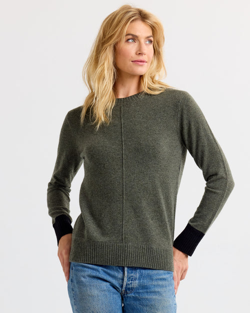 Three-quarter view image of a woman wearing a dark olive cashmere sweater with black cuffs and blue jeans, hands on hips