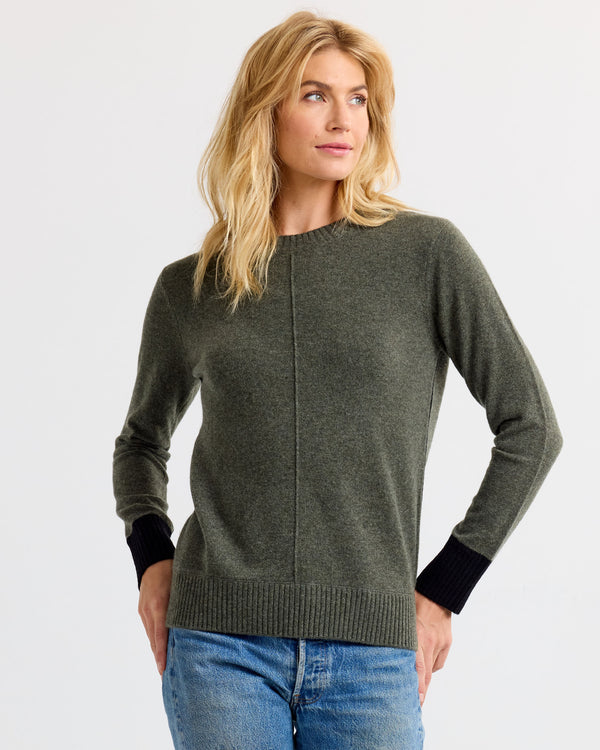 Three-quarter view image of a woman wearing a dark olive cashmere sweater with black cuffs and blue jeans, hands on hips