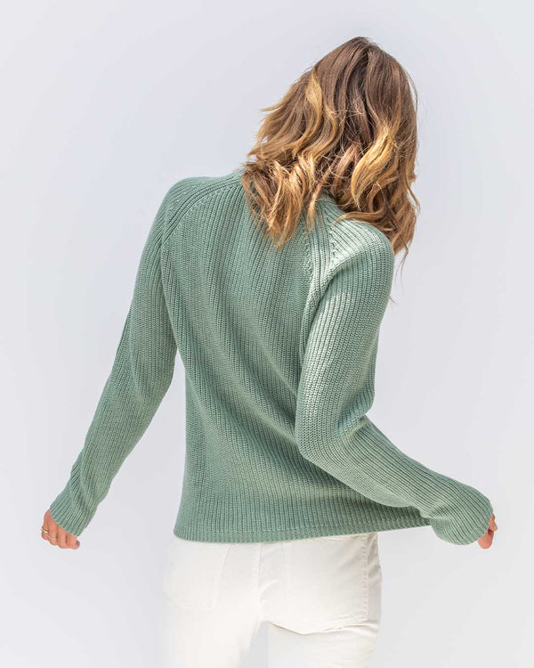 Woman wearing sage green ribbed cotton fisherman crew sweater, shown from back, paired with white pants against white