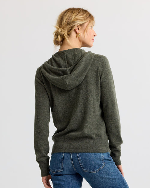 Woman wearing dark olive cashmere hoodie, back view, paired with blue jeans against plain background
