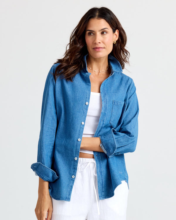 Woman wearing medium wash denim shirt over white top and white pants against plain background