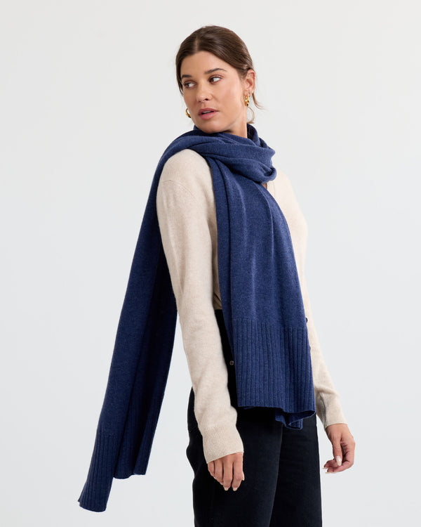 Side view image of a woman wearing an indigo cashmere scarf draped over a beige sweater against a plain background