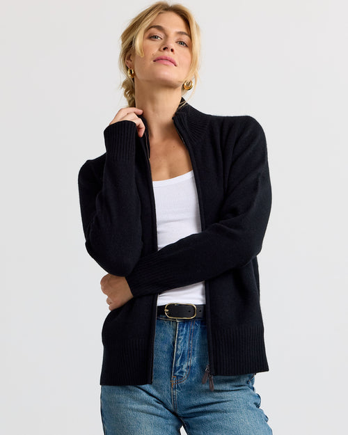 Front view image of a woman wearing black zip-up cashmere cardigan over white tank and blue jeans against plain background