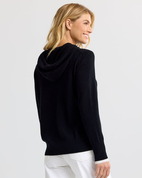 Woman wearing black cashmere hoodie with white cuffs, shown from back, smiling, white pants against plain background