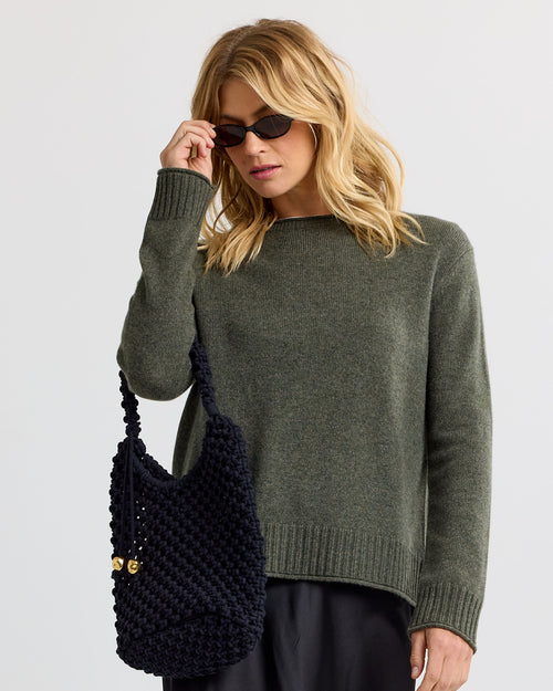 LIFESTYLE / EDITORIAL: Woman wearing dark olive cashmere sweater and black knit bag, adjusting sunglasses against white